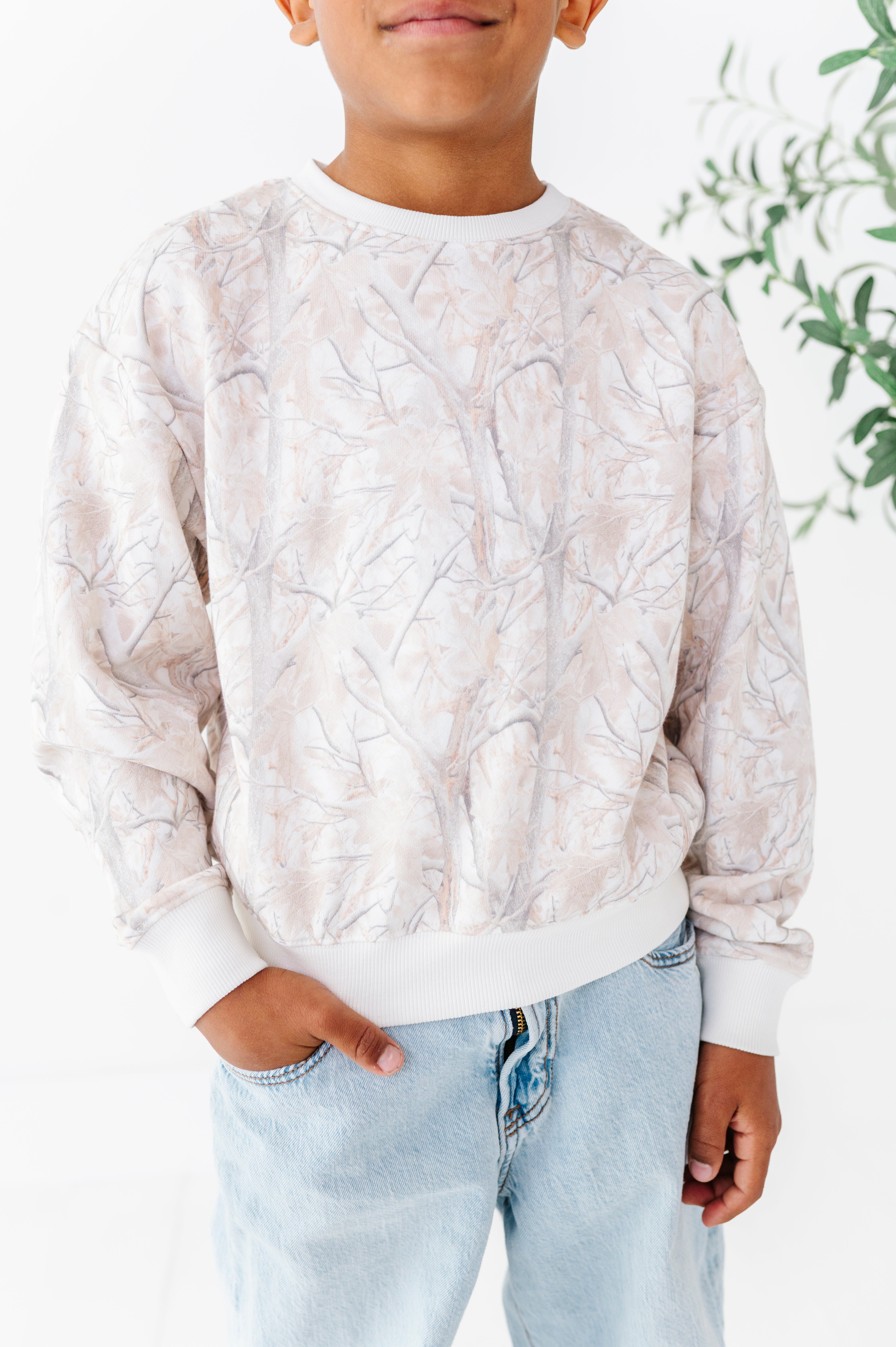 Child wearing a light-colored camo-patterned sweatshirt with light blue jeans against a white background.